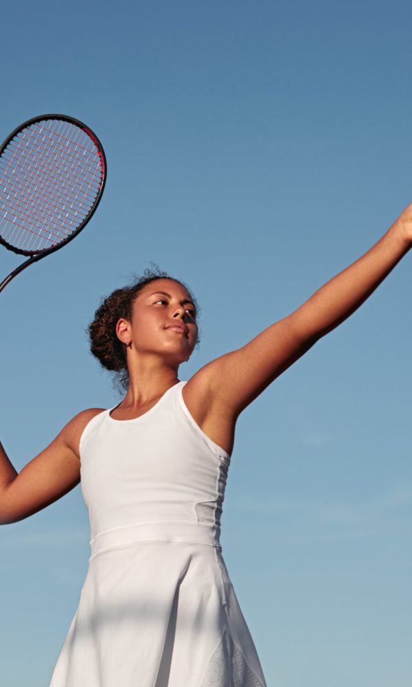 From below African American female athlete in white dress preparing to hit ball with racket against blue sky while playing tennis in daytime