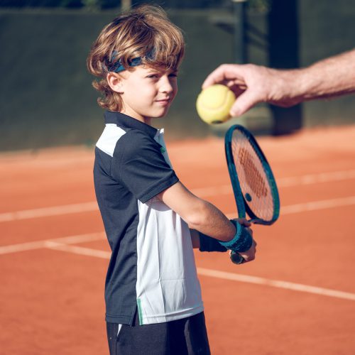 Side view of focused cute preteen boy in sportswear preparing backhand stroke with tennis racket on ball in hand of coach while training on hard court