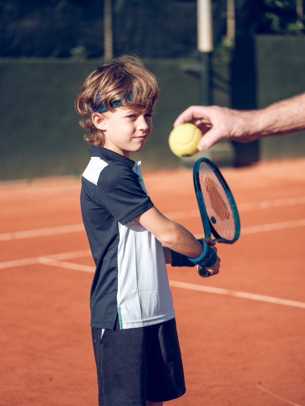 Side view of focused cute preteen boy in sportswear preparing backhand stroke with tennis racket on ball in hand of coach while training on hard court